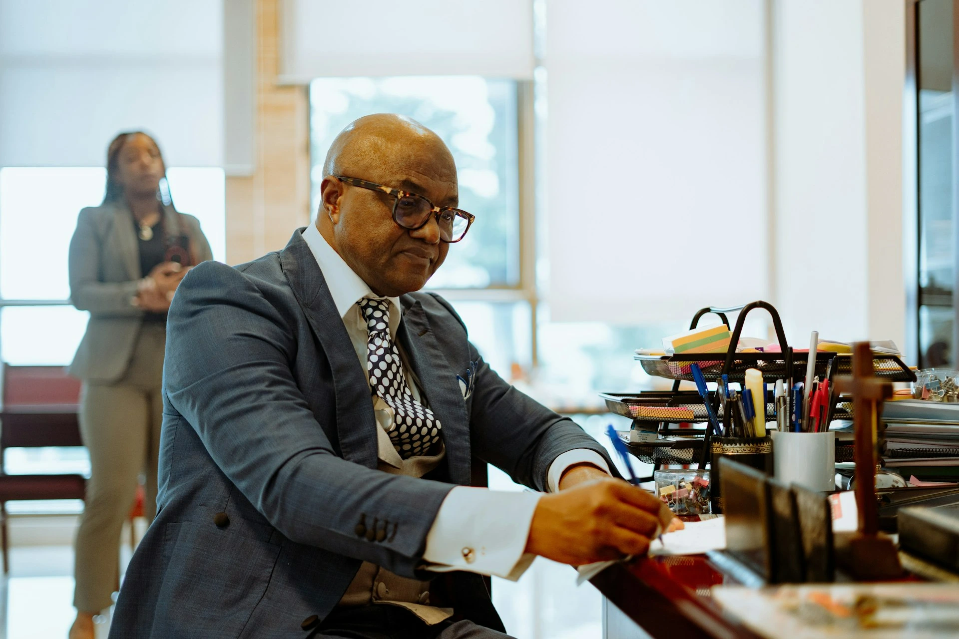 A man in a blue suit writing at a desk with a woman standing behind